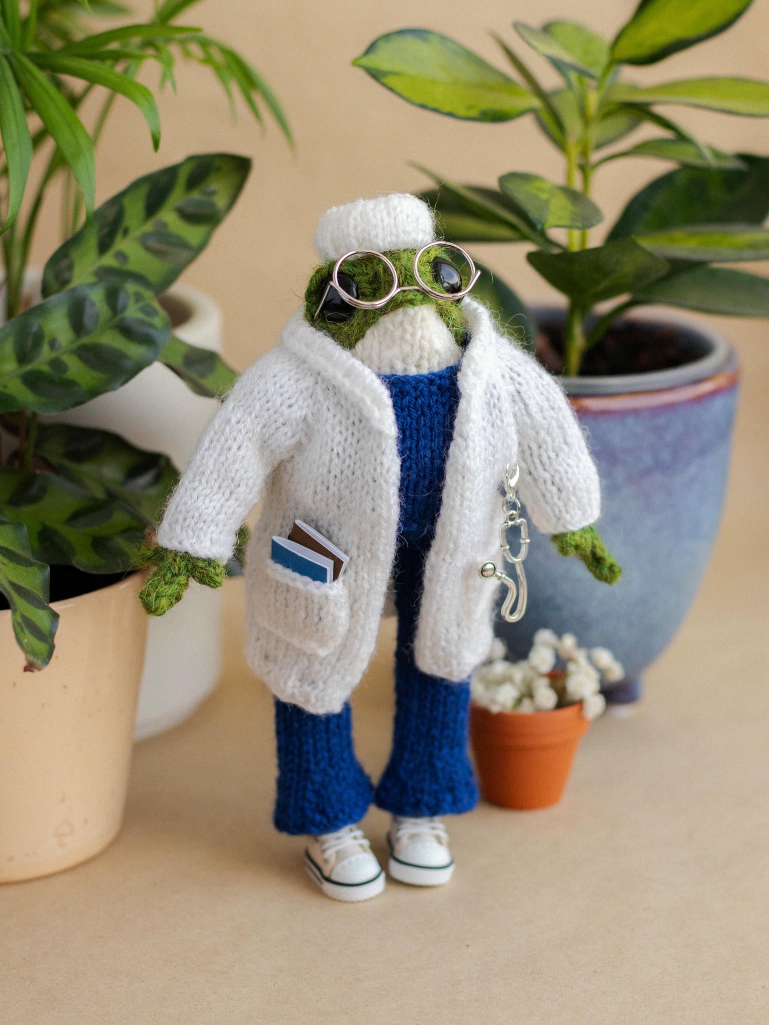 Knitted frog toy wearing a white coat and blue pants with plants in the background