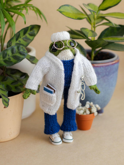 Knitted frog toy wearing a white coat and blue pants with plants in the background