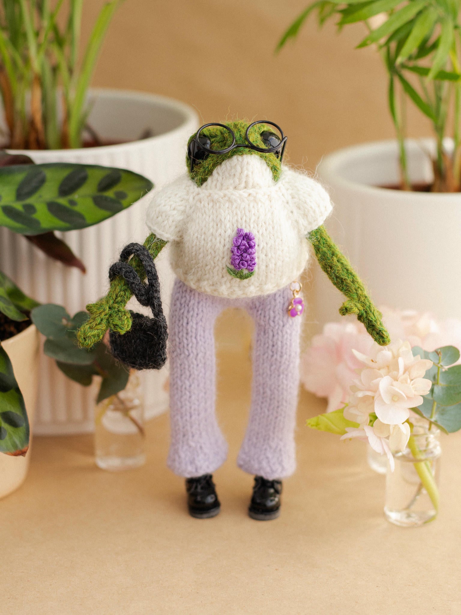 Knitted frog character wearing a sweater and pants, surrounded by plants and flowers.