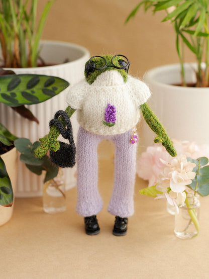 Knitted frog character wearing a sweater and pants, surrounded by plants and flowers.