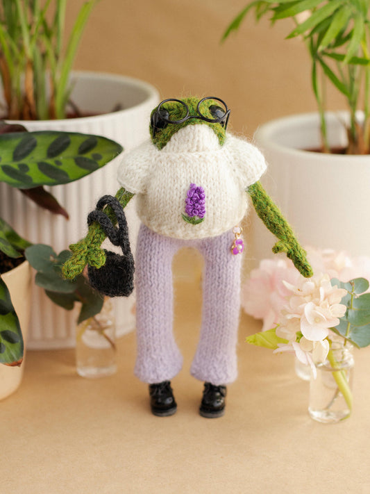 Knitted frog character wearing a sweater and pants, surrounded by plants and flowers.