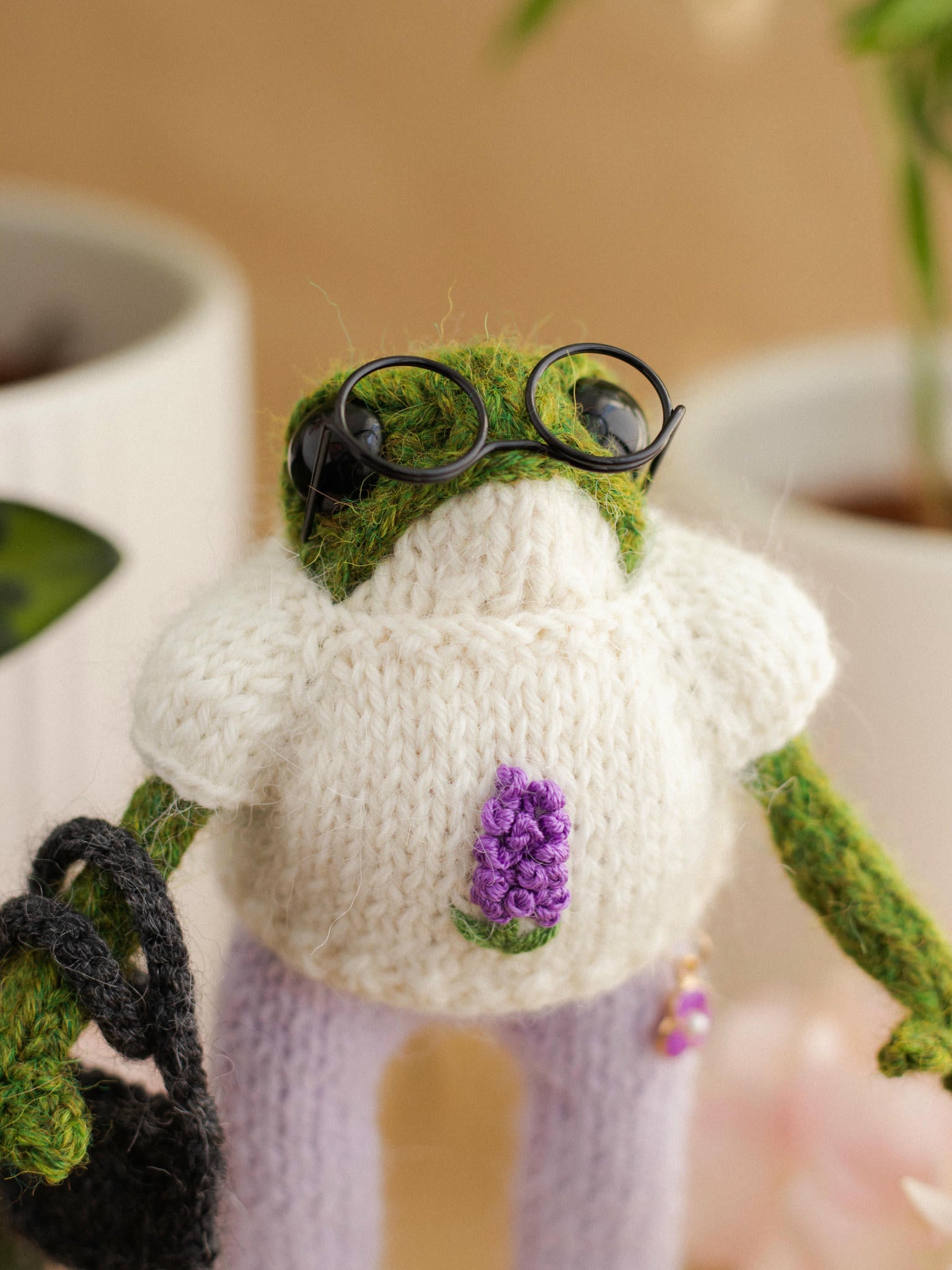 Knitted frog wearing glasses and a sweater with a flower, sitting on a surface.