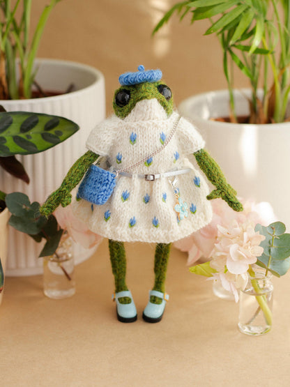 Frog toy dressed in a white outfit with blue accents, standing on a beige surface with plants in the background.