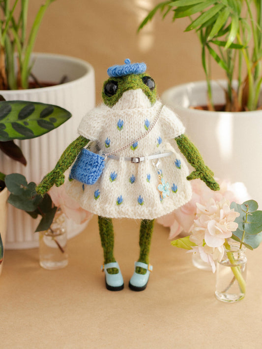 Frog toy dressed in a white outfit with blue accents, standing on a beige surface with plants in the background.